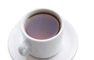 Tea with The Editor A top-down, high-angle view of a white ceramic teacup filled with dark amber tea or black coffee, resting on a matching round white saucer isolated on a plain background.