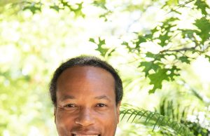 Fit and Functional Smiling man with long dreadlocks wearing a black shirt with a green and white "FF" logo, standing outdoors with green foliage in the background.