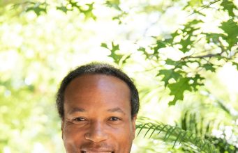 Fit and Functional Smiling man with long dreadlocks wearing a black shirt with a green and white "FF" logo, standing outdoors with green foliage in the background.