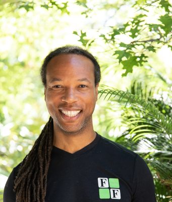Fit and Functional Smiling man with long dreadlocks wearing a black shirt with a green and white "FF" logo, standing outdoors with green foliage in the background.
