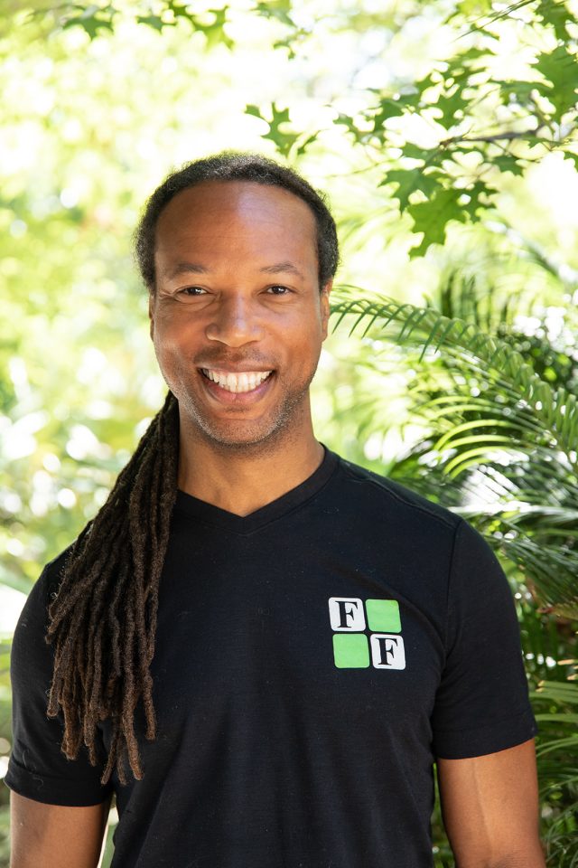 Fit & Functional Smiling man with long dreadlocks wearing a black shirt with a green and white "FF" logo, standing outdoors with green foliage in the background.