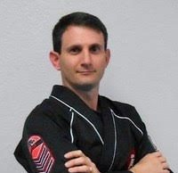 Kickstart Parenting A studio-style portrait of a man wearing a black martial arts uniform with arms crossed, standing against a plain light-colored background. The uniform includes visible rank insignia on the sleeve, and the subject is facing the camera with a composed expression.