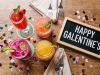 Celebrate Galentine’s Day An overhead photograph captures a cluster of four colorful cocktails arranged centrally on a wooden brunch table. Surrounding them is colorful heart-shaped confetti, two sets of folded tan linen napkins with forks, and a small, framed chalkboard sign that reads "HAPPY GALENTINE'S". The top-left drink is a tall glass of red mimosa with mint and berries. To its right is a round pink cocktail with a salted rim and basil. In the foreground is a bright pink dragon fruit cocktail with a lime wheel, and to its right is a short glass jar of orange-yellow juice with a bamboo pick. The scene is well-lit and festive.