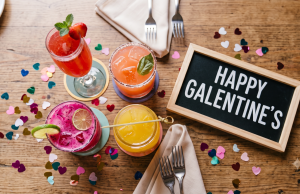 Celebrate Galentine’s Day An overhead photograph captures a cluster of four colorful cocktails arranged centrally on a wooden brunch table. Surrounding them is colorful heart-shaped confetti, two sets of folded tan linen napkins with forks, and a small, framed chalkboard sign that reads "HAPPY GALENTINE'S". The top-left drink is a tall glass of red mimosa with mint and berries. To its right is a round pink cocktail with a salted rim and basil. In the foreground is a bright pink dragon fruit cocktail with a lime wheel, and to its right is a short glass jar of orange-yellow juice with a bamboo pick. The scene is well-lit and festive.