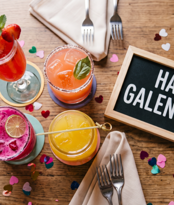 Celebrate Galentine’s Day An overhead photograph captures a cluster of four colorful cocktails arranged centrally on a wooden brunch table. Surrounding them is colorful heart-shaped confetti, two sets of folded tan linen napkins with forks, and a small, framed chalkboard sign that reads "HAPPY GALENTINE'S". The top-left drink is a tall glass of red mimosa with mint and berries. To its right is a round pink cocktail with a salted rim and basil. In the foreground is a bright pink dragon fruit cocktail with a lime wheel, and to its right is a short glass jar of orange-yellow juice with a bamboo pick. The scene is well-lit and festive.
