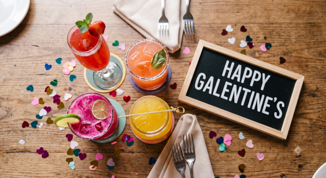 Galentines An overhead photograph captures a cluster of four colorful cocktails arranged centrally on a wooden brunch table. Surrounding them is colorful heart-shaped confetti, two sets of folded tan linen napkins with forks, and a small, framed chalkboard sign that reads "HAPPY GALENTINE'S". The top-left drink is a tall glass of red mimosa with mint and berries. To its right is a round pink cocktail with a salted rim and basil. In the foreground is a bright pink dragon fruit cocktail with a lime wheel, and to its right is a short glass jar of orange-yellow juice with a bamboo pick. The scene is well-lit and festive.