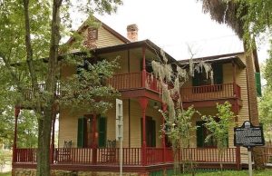 Walking through history at the Riley House The John G. Riley House, a historic two-story wood-frame home with wraparound porches, red railings, green shutters, and surrounding trees.