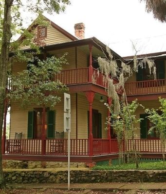 Walking through history at the Riley House The John G. Riley House, a historic two-story wood-frame home with wraparound porches, red railings, green shutters, and surrounding trees.
