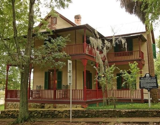 Walking through history at the Riley House The John G. Riley House, a historic two-story wood-frame home with wraparound porches, red railings, green shutters, and surrounding trees.