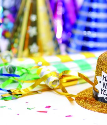 Rings in 2026 at Cascades Park Gold glitter top hat with a “Happy New Year!” label sits on a table surrounded by colorful party hats, streamers, and confetti, suggesting a New Year’s celebration.