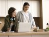 New Year’s resolutions to make kids sharper at home and smarter at school A woman and a teenage girl stand together in a bright kitchen, smiling as they look at a laptop on the counter. The teenager, wearing glasses and headphones around her neck, holds a pen and notepad, while the woman prepares food beside a bowl of salad ingredients.