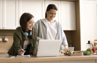 New Year’s resolutions to make kids sharper at home and smarter at school A woman and a teenage girl stand together in a bright kitchen, smiling as they look at a laptop on the counter. The teenager, wearing glasses and headphones around her neck, holds a pen and notepad, while the woman prepares food beside a bowl of salad ingredients.