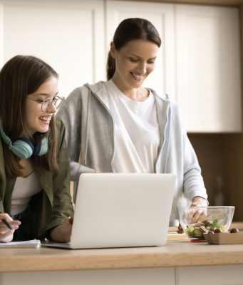 New Year’s resolutions to make kids sharper at home and smarter at school A woman and a teenage girl stand together in a bright kitchen, smiling as they look at a laptop on the counter. The teenager, wearing glasses and headphones around her neck, holds a pen and notepad, while the woman prepares food beside a bowl of salad ingredients.