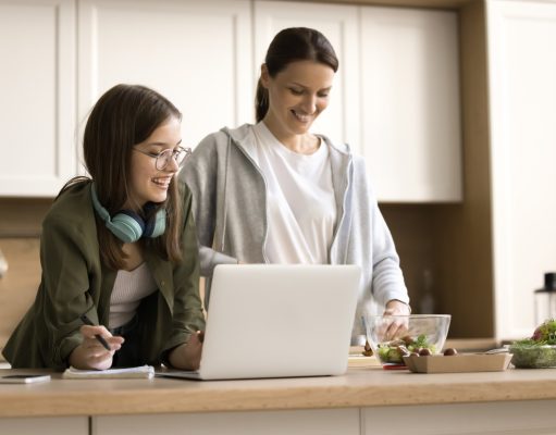 New Year’s resolutions to make kids sharper at home and smarter at school A woman and a teenage girl stand together in a bright kitchen, smiling as they look at a laptop on the counter. The teenager, wearing glasses and headphones around her neck, holds a pen and notepad, while the woman prepares food beside a bowl of salad ingredients.