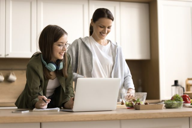 abaff1b1 A woman and a teenage girl stand together in a bright kitchen, smiling as they look at a laptop on the counter. The teenager, wearing glasses and headphones around her neck, holds a pen and notepad, while the woman prepares food beside a bowl of salad ingredients.