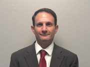 Kickstart Parenting Professional waist-up portrait of a man with short dark hair standing against a plain gray background. He is wearing a dark gray suit jacket, a white button-down collared shirt, and a textured red tie.
