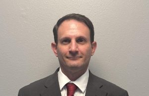 Kickstart Parenting Professional waist-up portrait of a man with short dark hair standing against a plain gray background. He is wearing a dark gray suit jacket, a white button-down collared shirt, and a textured red tie.