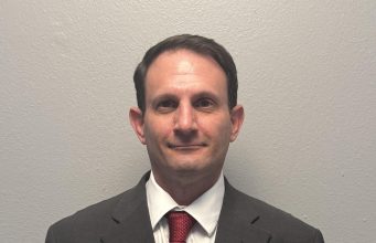 Kickstart Parenting Professional waist-up portrait of a man with short dark hair standing against a plain gray background. He is wearing a dark gray suit jacket, a white button-down collared shirt, and a textured red tie.