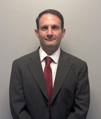 Kickstart Parenting Professional waist-up portrait of a man with short dark hair standing against a plain gray background. He is wearing a dark gray suit jacket, a white button-down collared shirt, and a textured red tie.