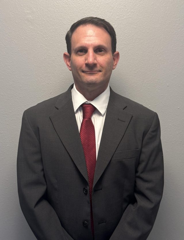 Professional waist-up portrait of a man with short dark hair standing against a plain gray background. He is wearing a dark gray suit jacket, a white button-down collared shirt, and a textured red tie.