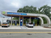 City opens first PowerTLH EV fast-charging station on Earth Day An outdoor electric vehicle charging station operated by the City of Tallahassee under a modern, curved canopy. A white Ford Mustang Mach-E and a white Chevrolet Bolt city utility vehicle are parked in charging bays. The canopy features digital signs displaying 'TALLAHASSEE SEE THE FUTURE TODAY' and 'PowerTLH' logos against a backdrop of trees and an overcast sky.