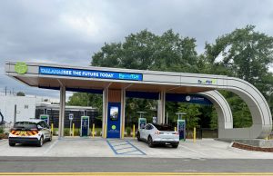 City opens first PowerTLH EV fast-charging station on Earth Day An outdoor electric vehicle charging station operated by the City of Tallahassee under a modern, curved canopy. A white Ford Mustang Mach-E and a white Chevrolet Bolt city utility vehicle are parked in charging bays. The canopy features digital signs displaying 'TALLAHASSEE SEE THE FUTURE TODAY' and 'PowerTLH' logos against a backdrop of trees and an overcast sky.