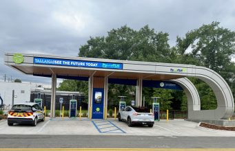 City opens first PowerTLH EV fast-charging station on Earth Day An outdoor electric vehicle charging station operated by the City of Tallahassee under a modern, curved canopy. A white Ford Mustang Mach-E and a white Chevrolet Bolt city utility vehicle are parked in charging bays. The canopy features digital signs displaying 'TALLAHASSEE SEE THE FUTURE TODAY' and 'PowerTLH' logos against a backdrop of trees and an overcast sky.