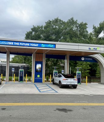 City opens first PowerTLH EV fast-charging station on Earth Day An outdoor electric vehicle charging station operated by the City of Tallahassee under a modern, curved canopy. A white Ford Mustang Mach-E and a white Chevrolet Bolt city utility vehicle are parked in charging bays. The canopy features digital signs displaying 'TALLAHASSEE SEE THE FUTURE TODAY' and 'PowerTLH' logos against a backdrop of trees and an overcast sky.