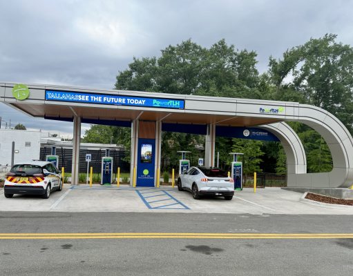 City opens first PowerTLH EV fast-charging station on Earth Day An outdoor electric vehicle charging station operated by the City of Tallahassee under a modern, curved canopy. A white Ford Mustang Mach-E and a white Chevrolet Bolt city utility vehicle are parked in charging bays. The canopy features digital signs displaying 'TALLAHASSEE SEE THE FUTURE TODAY' and 'PowerTLH' logos against a backdrop of trees and an overcast sky.
