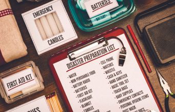 City of Tallahassee to host hurricane preparedness workshop in June A flat-lay view of emergency supplies arranged on a wooden table around a red clipboard with a 'Disaster Preparation List.' The checklist includes items like water, non-perishable food, a battery radio, batteries, a first aid kit, and matches. Surrounding the clipboard are physical prep items including boxes labeled 'Emergency Candles,' 'First Aid Kit,' and 'Matches,' alongside a clear container of batteries, a radio, a whistle, a pen, and a pair of pliers.