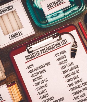 City of Tallahassee to host hurricane preparedness workshop in June A flat-lay view of emergency supplies arranged on a wooden table around a red clipboard with a 'Disaster Preparation List.' The checklist includes items like water, non-perishable food, a battery radio, batteries, a first aid kit, and matches. Surrounding the clipboard are physical prep items including boxes labeled 'Emergency Candles,' 'First Aid Kit,' and 'Matches,' alongside a clear container of batteries, a radio, a whistle, a pen, and a pair of pliers.