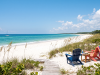 Share your “Summer in Bradfordville” photos A daytime photograph looking toward a pristine, deserted white sand beach and turquoise ocean under a bright blue sky with scattered clouds. The foreground consists of natural coastal dunes covered in sea oats and native scrub. A weathered wooden plank walkway leads from the vegetation onto the sand. In the mid-ground, a dark blue Adirondack chair and a coral-pink Adirondack chair face the water. A small wooden table between them holds a water bottle and sunglasses. The pink chair has a brightly striped beach towel draped over it. Far in the distance, a small white sailboat is anchored on the calm water. The composition is empty of people, emphasizing natural beauty and solitude.