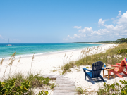 Share your “Summer in Bradfordville” photos A daytime photograph looking toward a pristine, deserted white sand beach and turquoise ocean under a bright blue sky with scattered clouds. The foreground consists of natural coastal dunes covered in sea oats and native scrub. A weathered wooden plank walkway leads from the vegetation onto the sand. In the mid-ground, a dark blue Adirondack chair and a coral-pink Adirondack chair face the water. A small wooden table between them holds a water bottle and sunglasses. The pink chair has a brightly striped beach towel draped over it. Far in the distance, a small white sailboat is anchored on the calm water. The composition is empty of people, emphasizing natural beauty and solitude.
