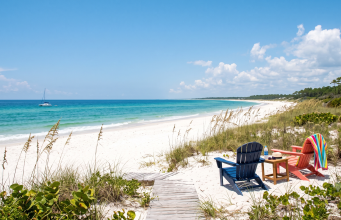 Share your “Summer in Bradfordville” photos A daytime photograph looking toward a pristine, deserted white sand beach and turquoise ocean under a bright blue sky with scattered clouds. The foreground consists of natural coastal dunes covered in sea oats and native scrub. A weathered wooden plank walkway leads from the vegetation onto the sand. In the mid-ground, a dark blue Adirondack chair and a coral-pink Adirondack chair face the water. A small wooden table between them holds a water bottle and sunglasses. The pink chair has a brightly striped beach towel draped over it. Far in the distance, a small white sailboat is anchored on the calm water. The composition is empty of people, emphasizing natural beauty and solitude.
