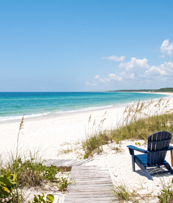 Share your “Summer in Bradfordville” photos A daytime photograph looking toward a pristine, deserted white sand beach and turquoise ocean under a bright blue sky with scattered clouds. The foreground consists of natural coastal dunes covered in sea oats and native scrub. A weathered wooden plank walkway leads from the vegetation onto the sand. In the mid-ground, a dark blue Adirondack chair and a coral-pink Adirondack chair face the water. A small wooden table between them holds a water bottle and sunglasses. The pink chair has a brightly striped beach towel draped over it. Far in the distance, a small white sailboat is anchored on the calm water. The composition is empty of people, emphasizing natural beauty and solitude.