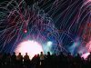 Celebrate America returns to Tom Brown Park for nation’s 250th birthday A vibrant nighttime photograph showing a silhouette of a crowd of spectators looking up at a massive, colorful fireworks display. Bright pink, blue, and green light trails arc across the black sky above a brilliant white burst of light at the center.