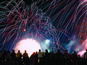 Celebrate America returns to Tom Brown Park for nation’s 250th birthday A vibrant nighttime photograph showing a silhouette of a crowd of spectators looking up at a massive, colorful fireworks display. Bright pink, blue, and green light trails arc across the black sky above a brilliant white burst of light at the center.