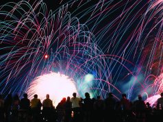 Celebrate America returns to Tom Brown Park for nation’s 250th birthday A vibrant nighttime photograph showing a silhouette of a crowd of spectators looking up at a massive, colorful fireworks display. Bright pink, blue, and green light trails arc across the black sky above a brilliant white burst of light at the center.