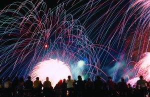 Celebrate America returns to Tom Brown Park for nation’s 250th birthday A vibrant nighttime photograph showing a silhouette of a crowd of spectators looking up at a massive, colorful fireworks display. Bright pink, blue, and green light trails arc across the black sky above a brilliant white burst of light at the center.