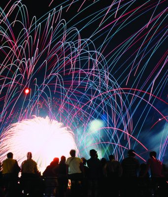 Celebrate America returns to Tom Brown Park for nation’s 250th birthday A vibrant nighttime photograph showing a silhouette of a crowd of spectators looking up at a massive, colorful fireworks display. Bright pink, blue, and green light trails arc across the black sky above a brilliant white burst of light at the center.