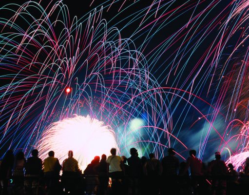 Celebrate America returns to Tom Brown Park for nation’s 250th birthday A vibrant nighttime photograph showing a silhouette of a crowd of spectators looking up at a massive, colorful fireworks display. Bright pink, blue, and green light trails arc across the black sky above a brilliant white burst of light at the center.
