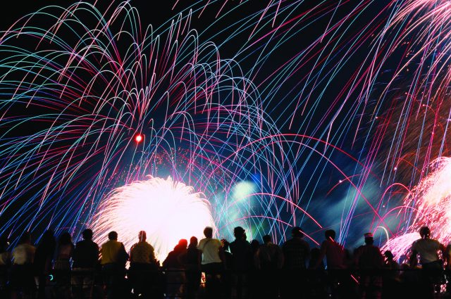 IMG_6625 A vibrant nighttime photograph showing a silhouette of a crowd of spectators looking up at a massive, colorful fireworks display. Bright pink, blue, and green light trails arc across the black sky above a brilliant white burst of light at the center.