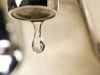 Annual report confirms Tallahassee drinking water meets all quality standards An extreme close-up photograph of a single, clear droplet of water hanging from the aerator tip of a chrome faucet just before falling. The background is softly blurred with warm, neutral tones.
