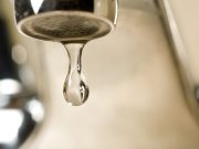 Annual report confirms Tallahassee drinking water meets all quality standards An extreme close-up photograph of a single, clear droplet of water hanging from the aerator tip of a chrome faucet just before falling. The background is softly blurred with warm, neutral tones.