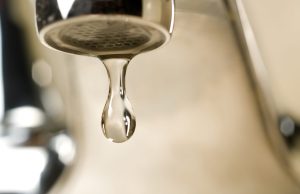 Annual report confirms Tallahassee drinking water meets all quality standards An extreme close-up photograph of a single, clear droplet of water hanging from the aerator tip of a chrome faucet just before falling. The background is softly blurred with warm, neutral tones.