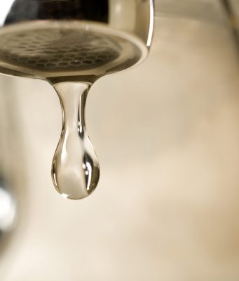 Annual report confirms Tallahassee drinking water meets all quality standards An extreme close-up photograph of a single, clear droplet of water hanging from the aerator tip of a chrome faucet just before falling. The background is softly blurred with warm, neutral tones.