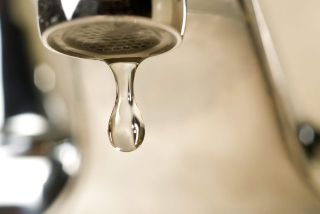 IMG_6630 An extreme close-up photograph of a single, clear droplet of water hanging from the aerator tip of a chrome faucet just before falling. The background is softly blurred with warm, neutral tones.