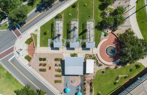 Remembering Smokey Hollow An aerial top-down view of Smokey Hollow Commemorative Park, featuring three open-frame house outline sculptures on green lawn, a brick circular plaza with a fountain, a central pavilion building, garden plots, and paved pedestrian walkways situated next to city streets.