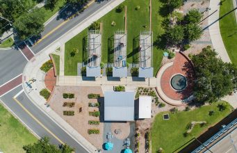 Remembering Smokey Hollow An aerial top-down view of Smokey Hollow Commemorative Park, featuring three open-frame house outline sculptures on green lawn, a brick circular plaza with a fountain, a central pavilion building, garden plots, and paved pedestrian walkways situated next to city streets.