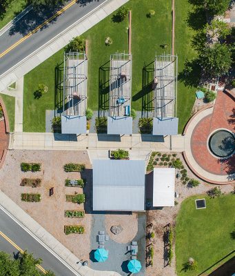 Remembering Smokey Hollow An aerial top-down view of Smokey Hollow Commemorative Park, featuring three open-frame house outline sculptures on green lawn, a brick circular plaza with a fountain, a central pavilion building, garden plots, and paved pedestrian walkways situated next to city streets.
